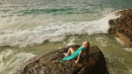 Fototapeta na wymiar Seductive long-haired woman sitting on rock of sea reef stone, stormy cloudy ocean. Woman in blue swimsuit dress tunic. Concept rest in sea, tropical resort coastline traveling tourism summer holidays