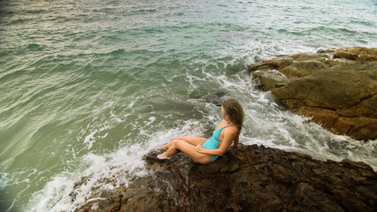 Handsome long-haired woman chill relax resting on rock of sea reef stone, stormy cloudy ocean. Woman in blue swimsuit dress tunic. Concept rest tropical resort coastline traveling outdoor tourism