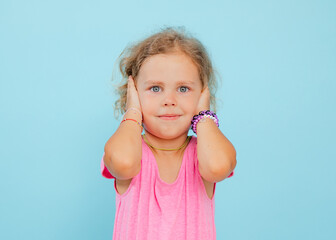 Charming little girl cover ears with hands on empty blue background, free copy space. Smiling child...
