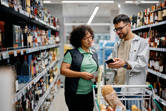 Young Man Using Smart Phone And Talking To Female Worker While Shopping In Supermarket.