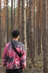 Alone young man in casual clothing and black hat with shoulder belt bag walking in a pine autumn forest. Rear view.