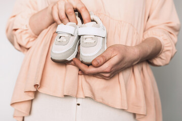 Pregnant woman in pink dress holding small shoes for unborn baby on belly, preparing go to maternity hospital for childbirth. Pregnancy, maternity, preparation, baby expectation concept. Copy space