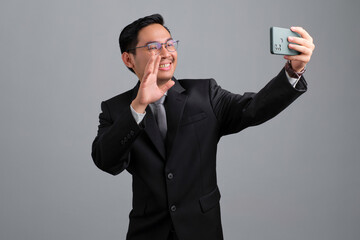 Portrait of cheerful handsome young businessman in formal suit and glasses waving hand while making video call on smartphone isolated on grey background