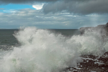Ocean wave hits rock coastline of Ireland creating splash of water. Wild Irish nature scene. Burren, county Clare. Cloudy sky.