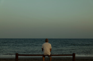 Fototapeta premium Adult man sitting on wooden fence looking at view of sea