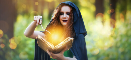 Young witch with spell book and magic wand in green forest