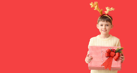 Funny little boy holding Christmas gift on red background with space for text