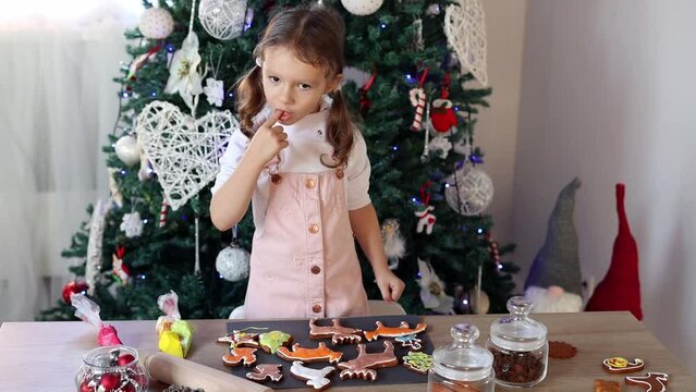 Happy little girl coloring Christmas gingerbread cookies at home in slow motion. Christmas decoration in the kitchen. Fir tree with fairy lights. The concept of the New Year and Christmas