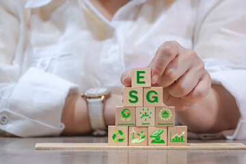 Businesswoman hand holding wooden cube with "ESG" text and icons on office desk, ESG concept of environmental, social and governance. Sustainable and ethical business