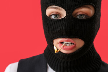 Portrait of young woman in balaclava with burning match in mouth on red background