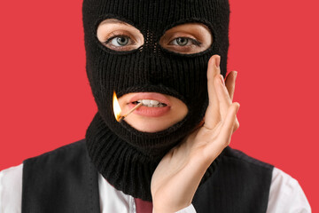 Portrait of young woman in balaclava with burning match in mouth on red background