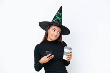 Young caucasian woman costume as witch isolated on white background holding coffee to take away and a mobile while thinking something