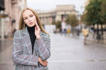 Fototapeta premium Portrait of a young and elegant Caucasian blonde girl in autumn in the city. The concept of femininity and style. A pretty girl is happy to pose in the city on the street in a coat.