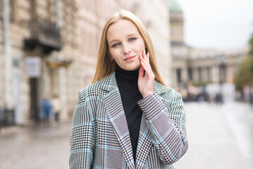 Fototapeta premium Portrait of a young and elegant Caucasian blonde girl in autumn in the city. The concept of femininity and style. A pretty girl is happy to pose in the city on the street in a coat.