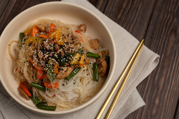 Funchoza work with chicken, carrots, sweet peppers and beans in a deep beige plate, metal sticks on a napkin, open space, flat lay