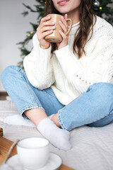 A girl in a white sweater, jeans and socks sits on the bed with a cup of hot drink in her hands and dreams. Behind the girl is a Christmas tree
