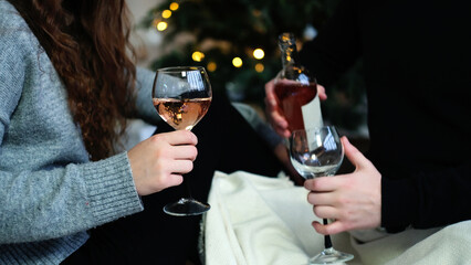 A loving couple sits on the floor covered with a blanket. A man pours wine into glasses. Christmas atmosphere