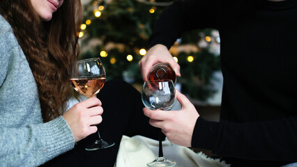 A loving couple sits on the floor covered with a blanket. A man pours wine into glasses. Christmas...