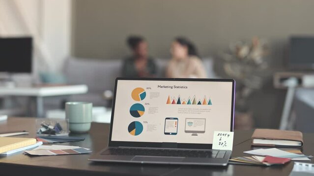 Slide with marketing statistics on laptop screen at office desk of marketing specialist. Two multiethnic female colleagues discussing new project on sofa in blurred background