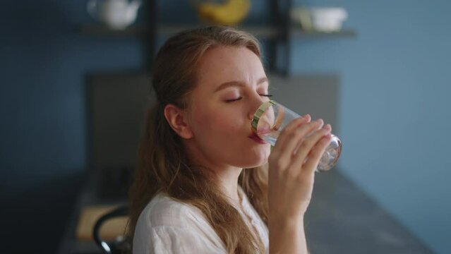 Thirsty smiling young woman standing alone in domestic kitchen drinking water from glass. Girl start new day with healthy life habit, sipping clean mineral natural still water close up view.