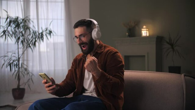 Young man celebration success using smartphone siting at couch.Bearded guy wear headphones looking at phone reading great news.Male winning lottery online cheering positive emotion. 
