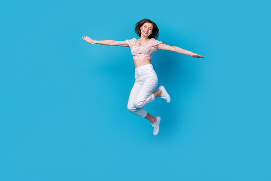 Photo of cute positive lady dressed trendy outfit jumping showing arm imagine can fly empty space isolated on blue color background