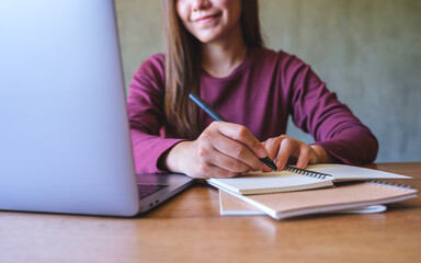 Closeup image of a young woman writing on a notebook while working on laptop computer