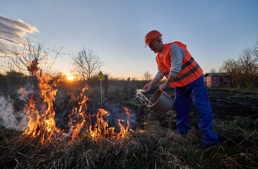 Fireman ecologist extinguishing fire in field with warning sign with exclamation mark end evening...