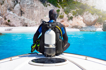 A scuba diver with his equipment sits on a bow of a boat in front of a beautiful beach with...