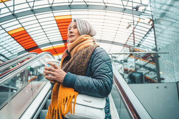 Senior woman at the station on escalator with coffee cup