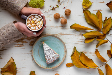 Top view of female hands holding marshmallows on shabby table with piece of cake, walnuts and scattered leaves