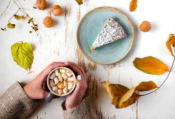 Top view of female hands holding marshmallows on shabby table with piece of cake, walnuts and scattered leaves