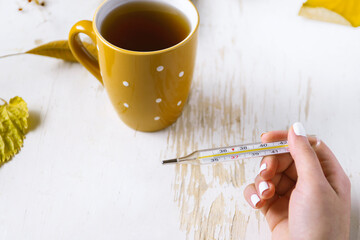Top view of female hands holding cup of tea and thermometer on shabby table with yellow scattered...