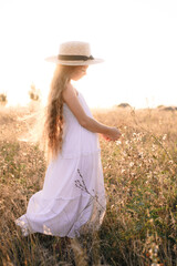 Cute little girl with blond long hair in a summer field at sunset with a white dress with a straw hat