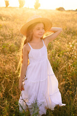 Cute little girl with blond long hair in a summer field at sunset with a white dress with a straw hat