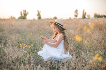 Fototapeta na wymiar Cute little girl with blond long hair in a summer field at sunset with a white dress with a straw hat