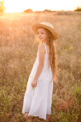 Cute little girl with blond long hair in a summer field at sunset with a white dress with a straw hat