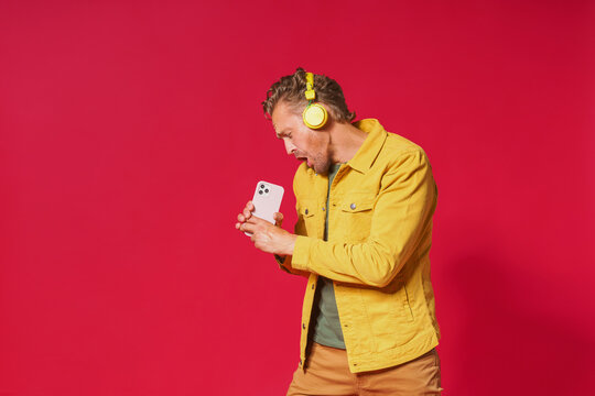 Joyful man sing while recording his voice using smartphone. Singing handsome man enjoying his favorite song using phone and wireless headphones wearing jeans yellow jacket isolated on red background