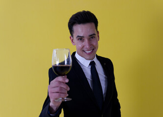 Celebration, dinner, elegance, party! A smiling handsome guy dressed in formal suit holding a wine glass while looking at camera ahead a yellow background