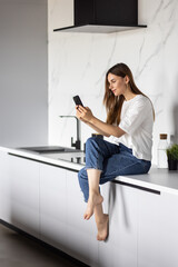 Young woman uses smartphone in the kitchen with laptop on the counter top.