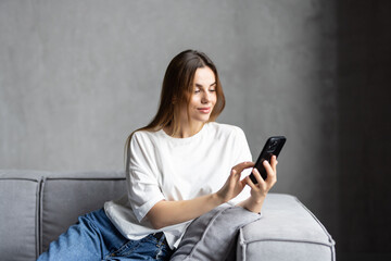 Naklejka premium Happy young woman using mobile phone while sitting a couch at home