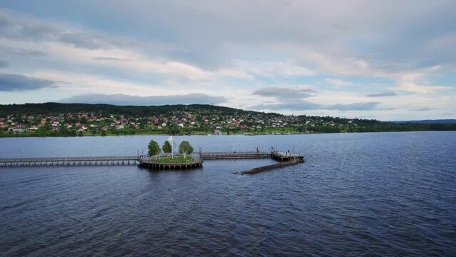 Aerial orbit shot. A pier at Lake Siljan in Sweden, people walk on the promenade. View of the town of Rättvik against the mountain and a harbor with sailing boats. Cloudy during sunset