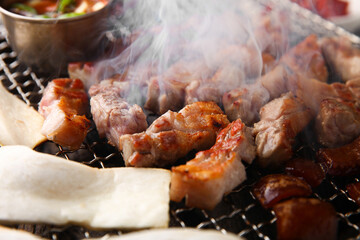 Grilled pork belly on a grill and set up a table with side dishes 그릴 위 노릇노릇하게 삼겹살을 굽고 있고 반찬이 세팅되어 있는 한상차림