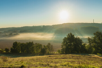 A view over a valley in the rolling hill landscape outside Maastricht, where the valley is covered with layers of fog and creating a nice setting with the sun beams through the mist over the fields