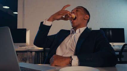 African american businessman eating donut near blurred laptop in office in evening.