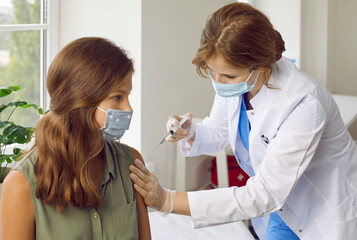 Doctor giving shot to child in face mask. Female nurse in white coat and facemask holding syringe...