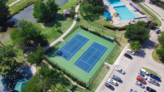 Aerial view large tennis court and swimming pool at community recreational center with trails and pond near Dallas, Texas, USA. Green park surrounded by mature trees, busy parking lots and outdoors