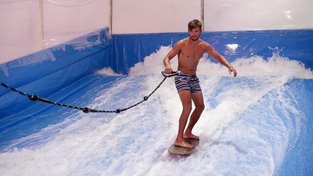 Attractive Man riding surf board on wave simulator indoors. Young surfer during training on generated waves. Water sport activity. Surfer guy enjoy surfing on indoor surf imitator. Surfing education