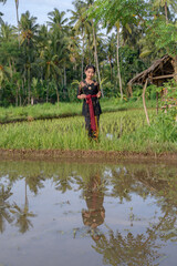 Naklejka na ściany i meble Balinese girl in green rice fields dressed in traditional costume dress black color reflecting in the water.
