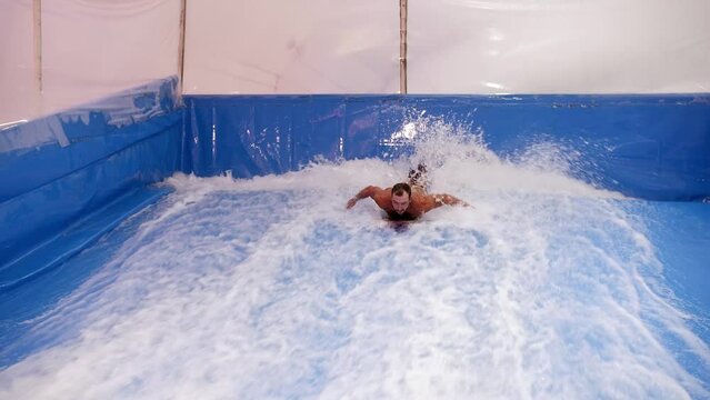 Attractive Man riding surf board on wave simulator indoors. Young surfer during training on generated waves. Water sport activity. Surfer guy enjoy surfing on indoor surf imitator. Surfing education